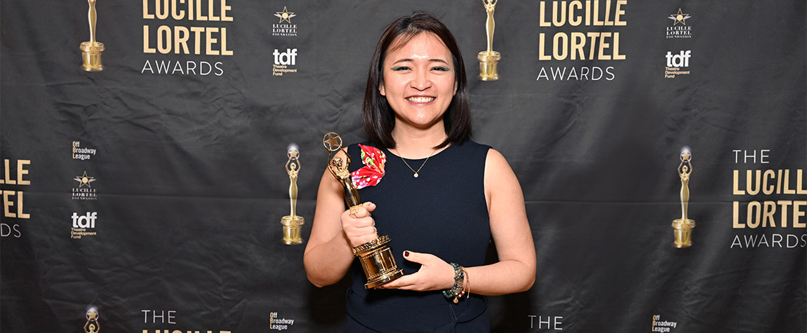 One person wearing a black dress holds an award statuette, in front of a photo backdrop with the words 